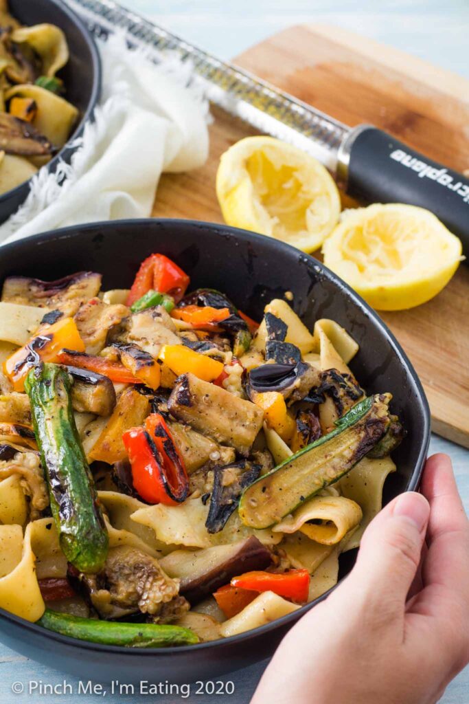 Hands holding a black bowl of pappardelle pasta with grilled vegetables. A cutting board with a lemon and zester are in the background.