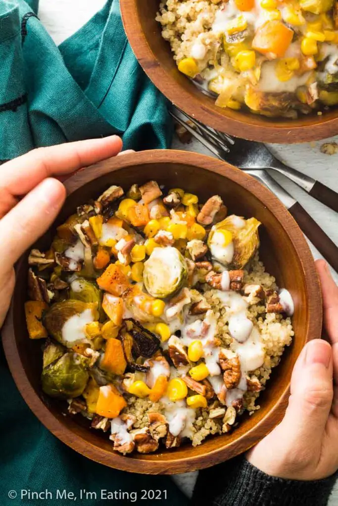 Hands holding a roasted vegetable quinoa harvest bowl in a wooden bowl with Brussels sprouts, quinoa, corn, sweet potatoes, shallots, pecans, and a creamy mozzarella sauce