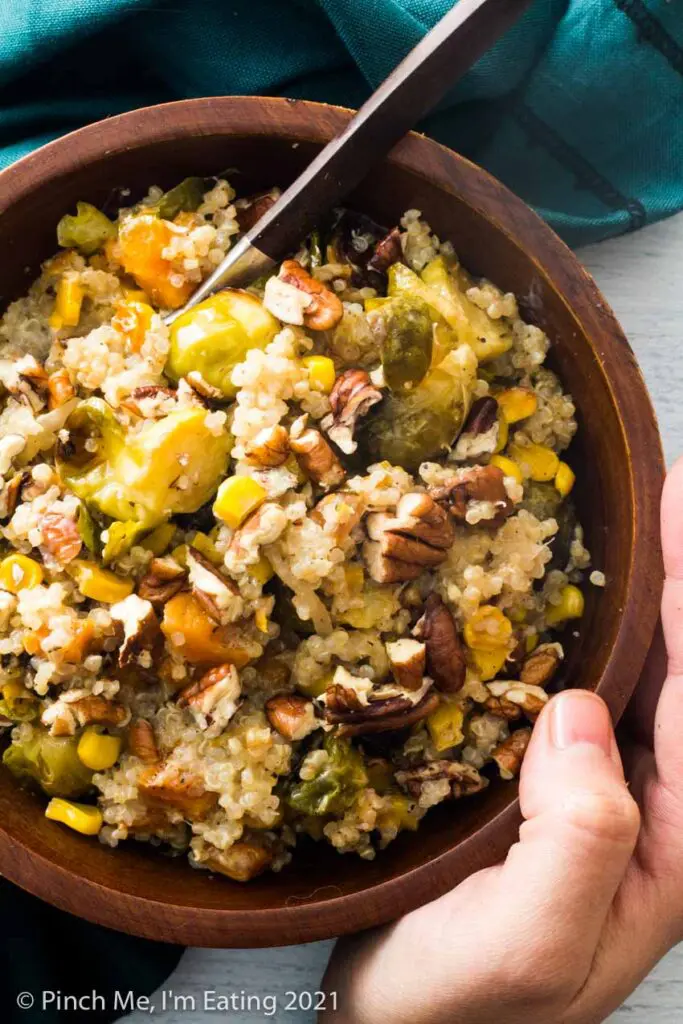 A hand holding a wooden bowl of Brussels sprouts, sweet potatoes, corn, shallots, quinoa, and pecans