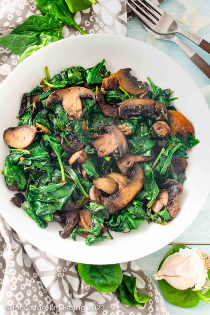 Overhead view of sautéed spinach and mushrooms in a white bowl with head of garlic and forks to the side