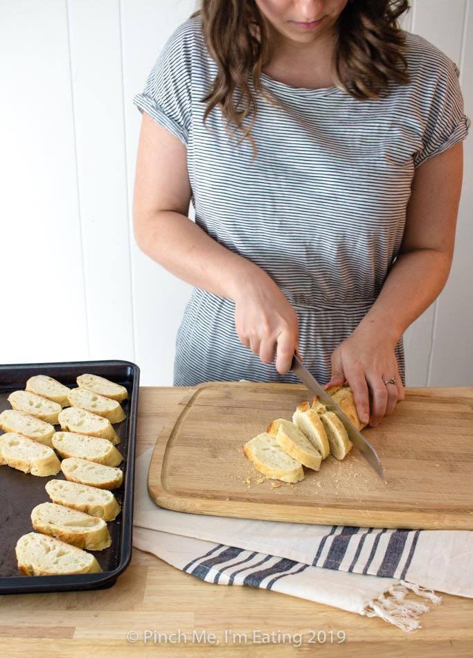 White woman in striped dress cutting a baguette on a wooden cutting board to make crostini