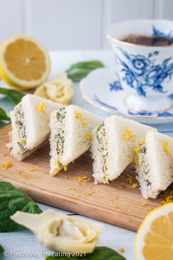 Three quarter view of artichoke spinach tea sandwiches arranged on a diagonal cutting board and topped with lemon zest. A blue and white teacup, half a lemon, artichoke, and spinach leaves are beside the board.