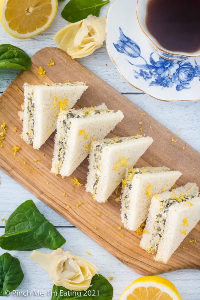 Overhead shot of artichoke spinach tea sandwiches arranged on a diagonal cutting board and topped with lemon zest. A blue and white teacup, half a lemon, artichoke, and spinach leaves are beside the board.