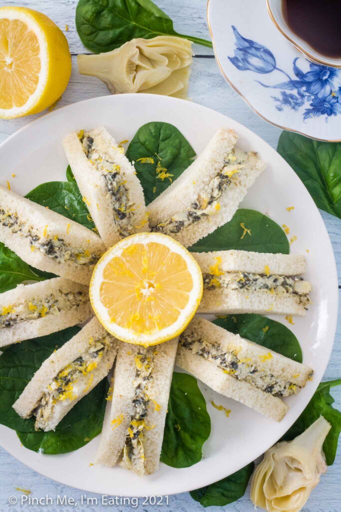 Overhead view of artichoke spinach tea sandwiches arranged on a white plate atop fresh spinach leaves, with half a lemon in the center of the plate. Blue and white teacup with tea is next to the plate.