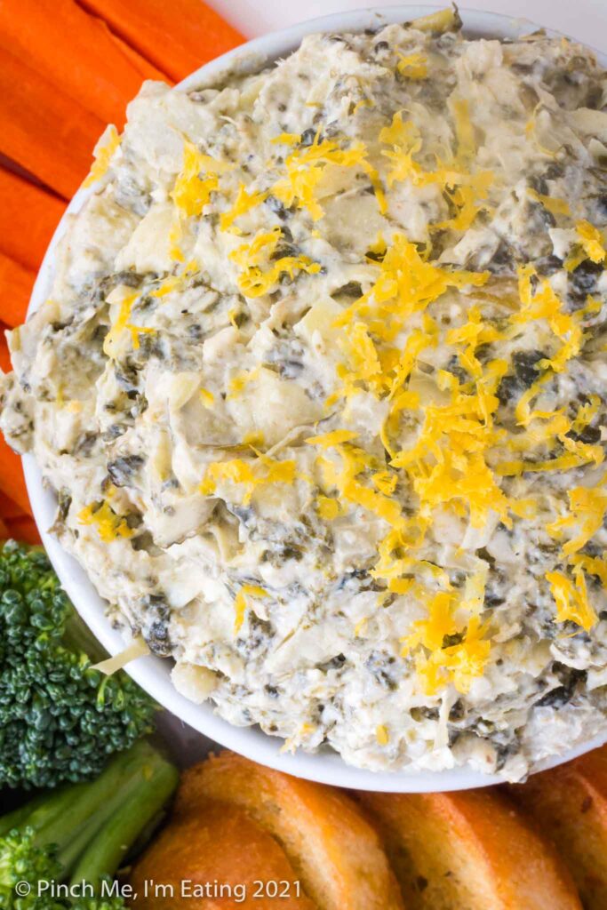 Overhead view of white bowl of cold artichoke spinach dip topped with lemon zest, surrounded by crostini and fresh vegetables