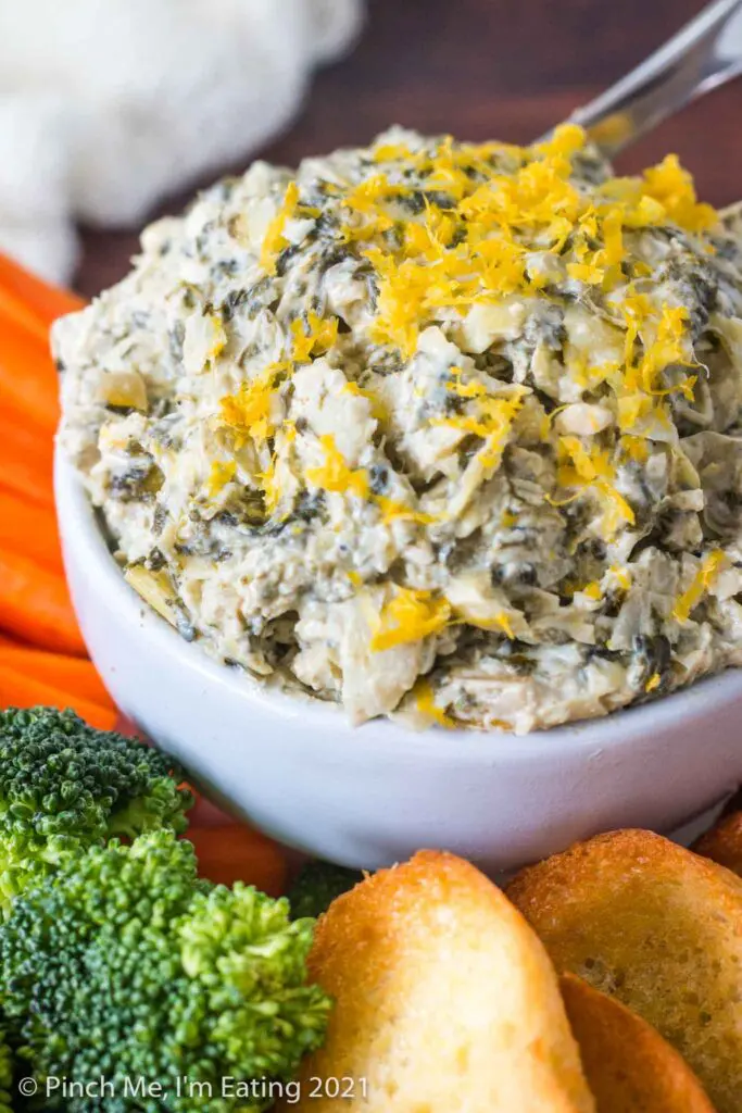 Three quarter view of white bowl of cold artichoke spinach dip topped with lemon zest, surrounded by crostini and fresh vegetables
