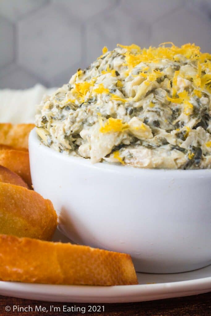 Heaping white bowl of cold artichoke spinach dip topped with lemon zest, surrounded by crostini