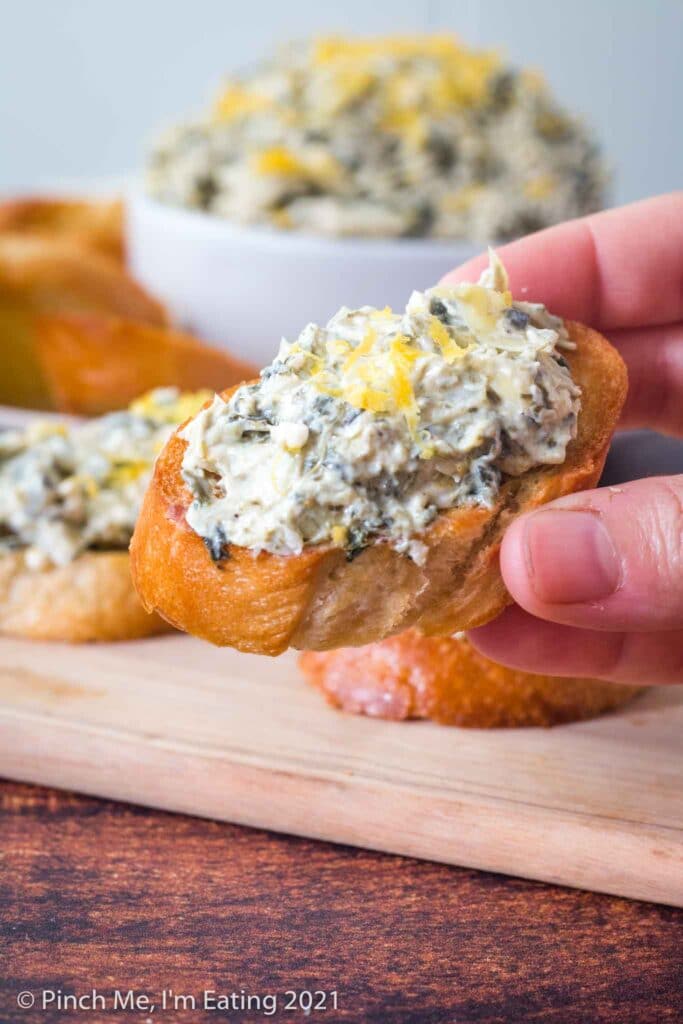 Hand holding crostini topped with cold artichoke spinach dip with lemon zest
