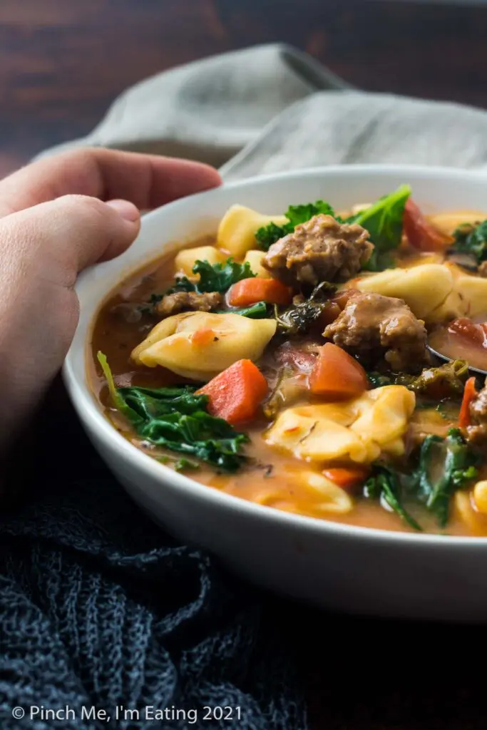 Moody shot of a hand holding a white bowl of Italian sausage soup with tortellini and kale