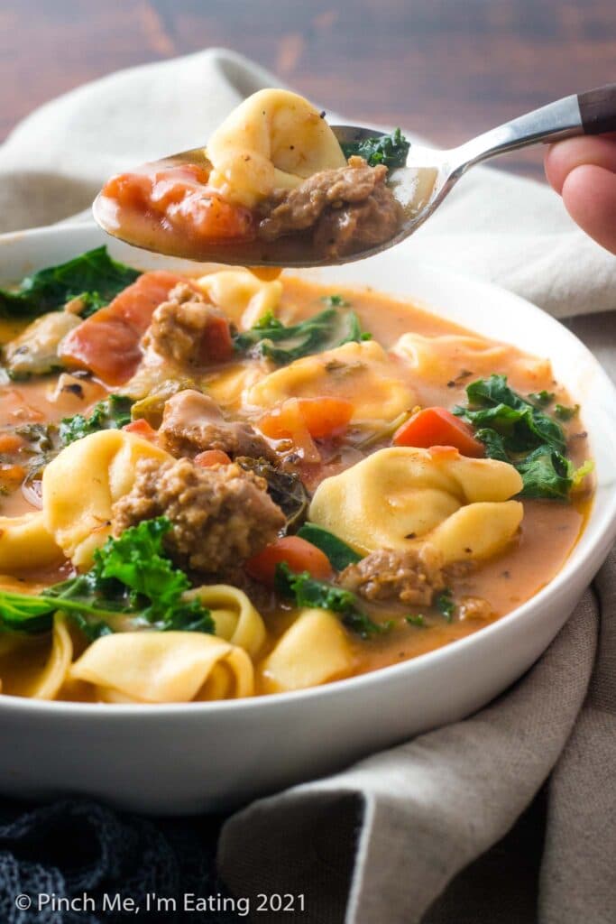 Three quarter view of a spoonful of Italian sausage soup with tortellini and kale over a white bowl of soup