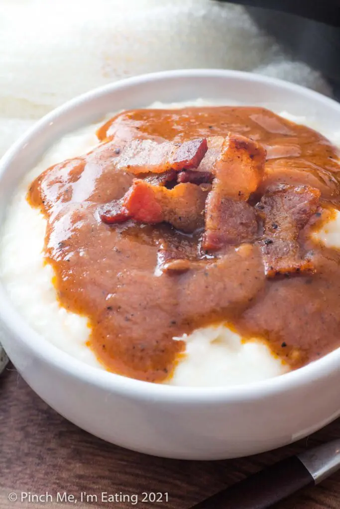 Close up view of grits in white bowl topped with bacon and Southern tomato gravy
