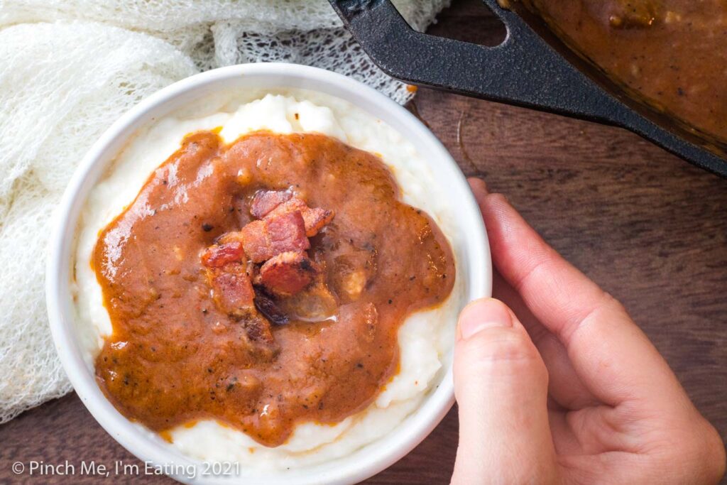 Overhead view of grits in white bowl topped with bacon and Southern tomato gravy, next to cast iron skillet