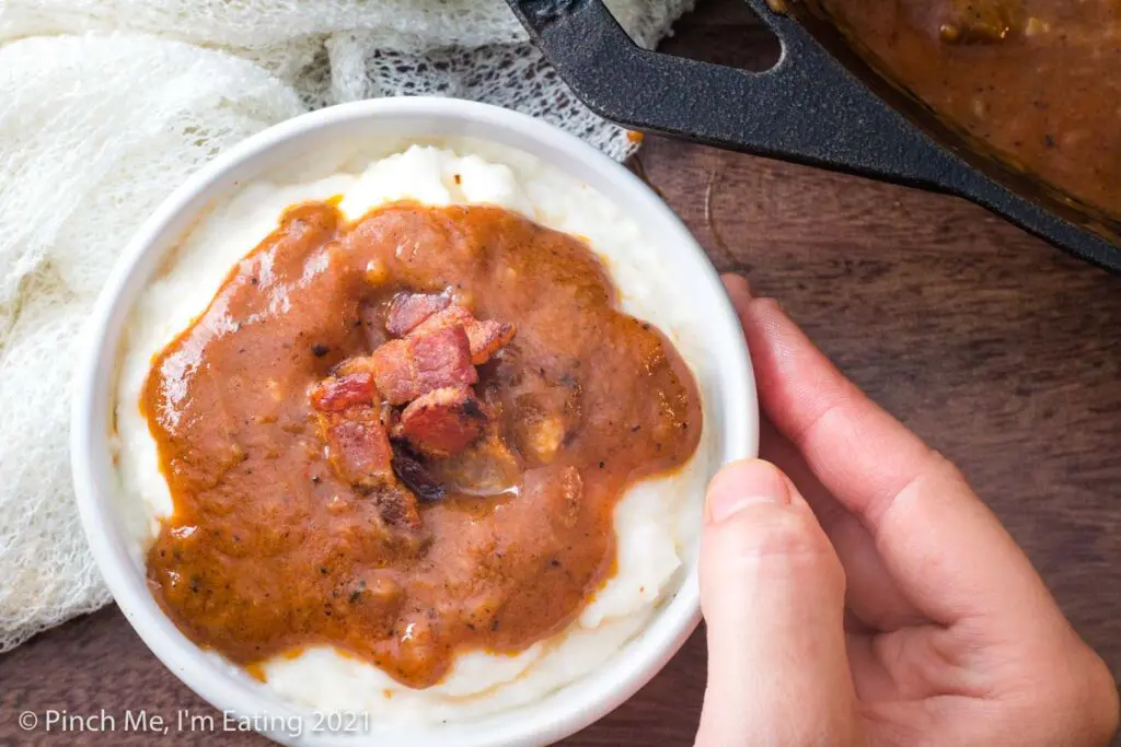 Overhead view of grits in white bowl topped with bacon and Southern tomato gravy, next to cast iron skillet