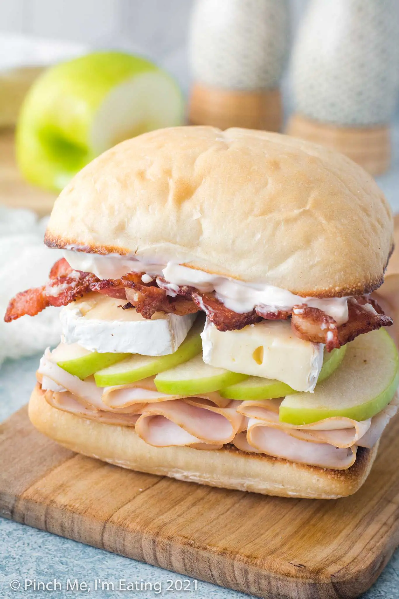 A sandwich sitting on a wooden cutting board is layered on a ciabatta roll with smoked deli turkey, green granny smith apple, brie cheese, bacon, and garlic mayo.