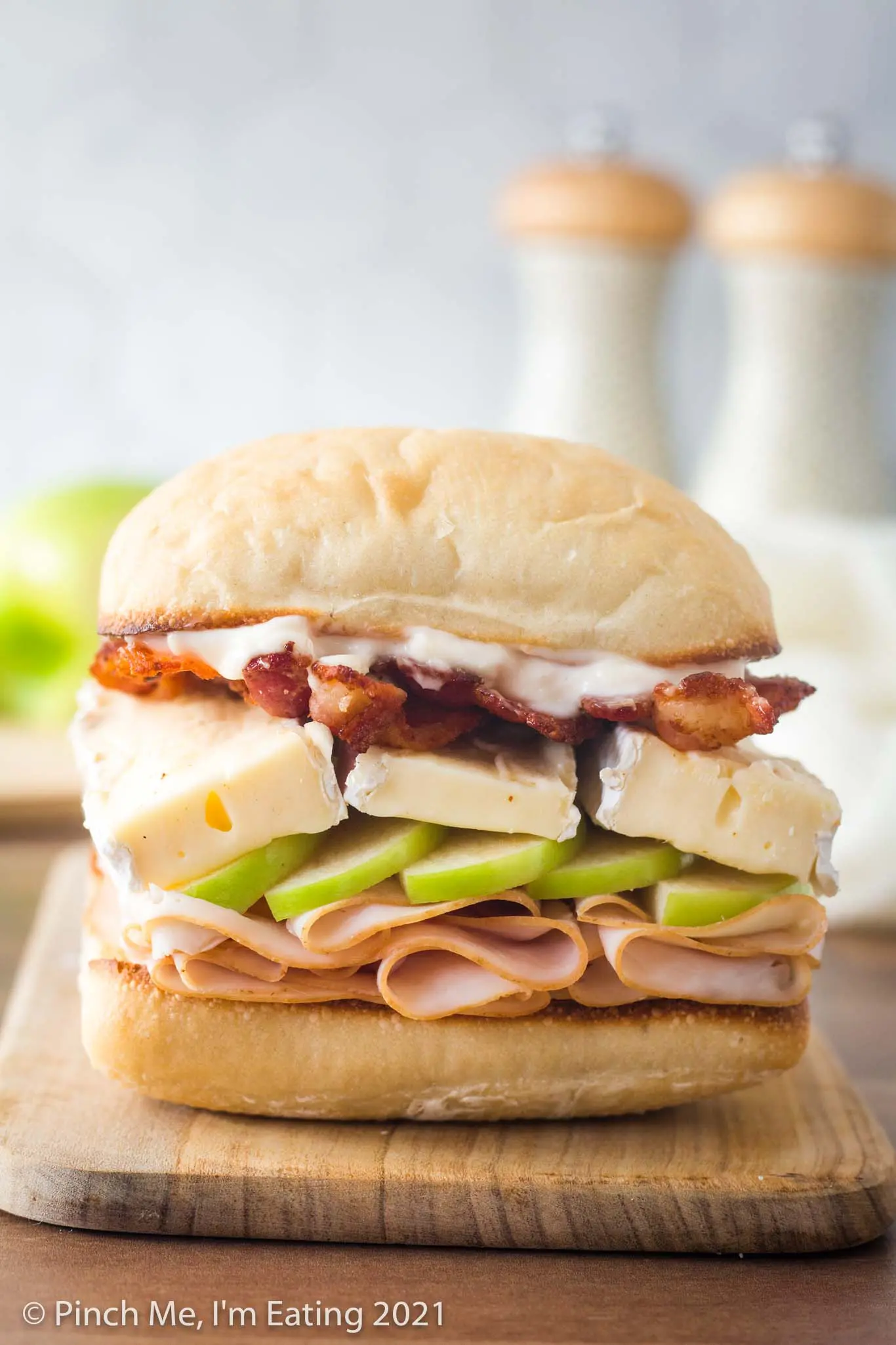 A sandwich sitting on a wooden cutting board is layered on a ciabatta roll with smoked deli turkey, green granny smith apple, brie cheese, bacon, and garlic mayo.
