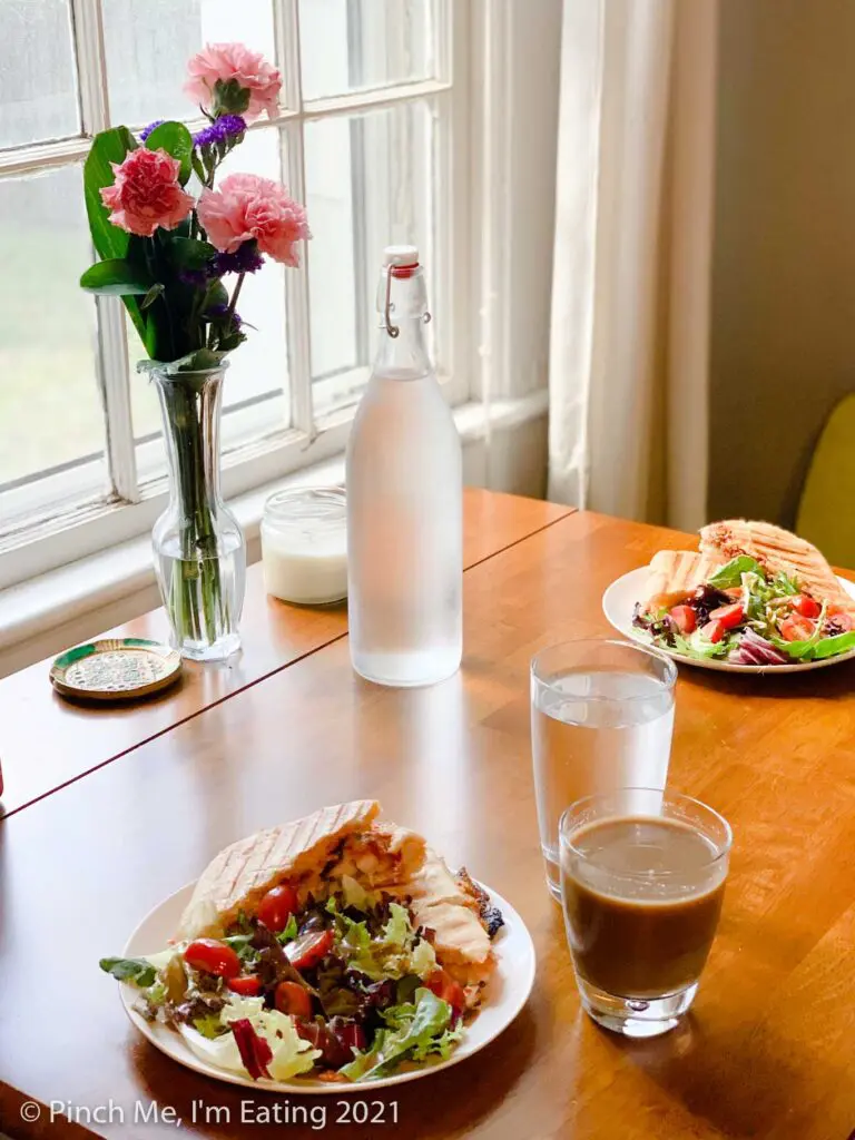 Cafe table in dining room next to window with fresh flowers and paninis on white plates with side salads. Water is in a glass water bottle.