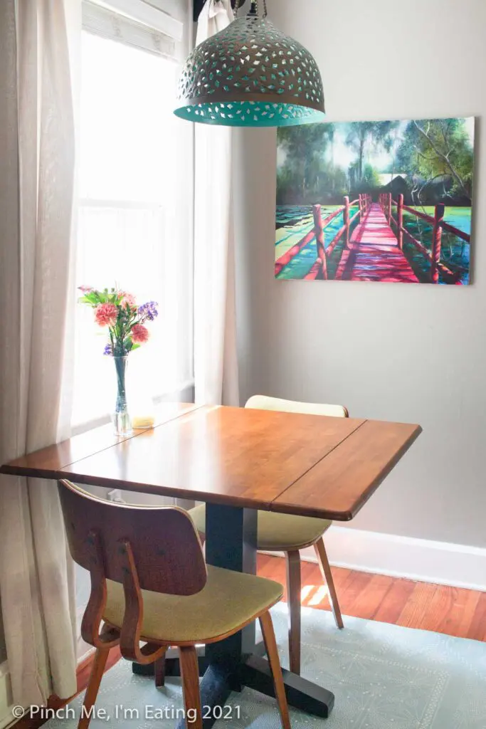 Cafe table in dining room next to window with pendant light and fresh flowers with art on the wall