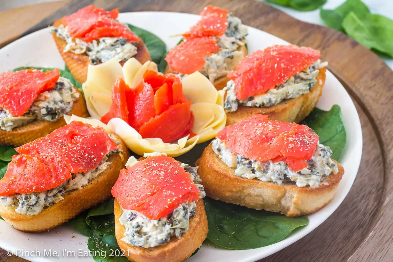 Artichoke spinach dip crostini with smoked salmon arranged in on a white plate around a flower made of artichoke heart and smoked salmon