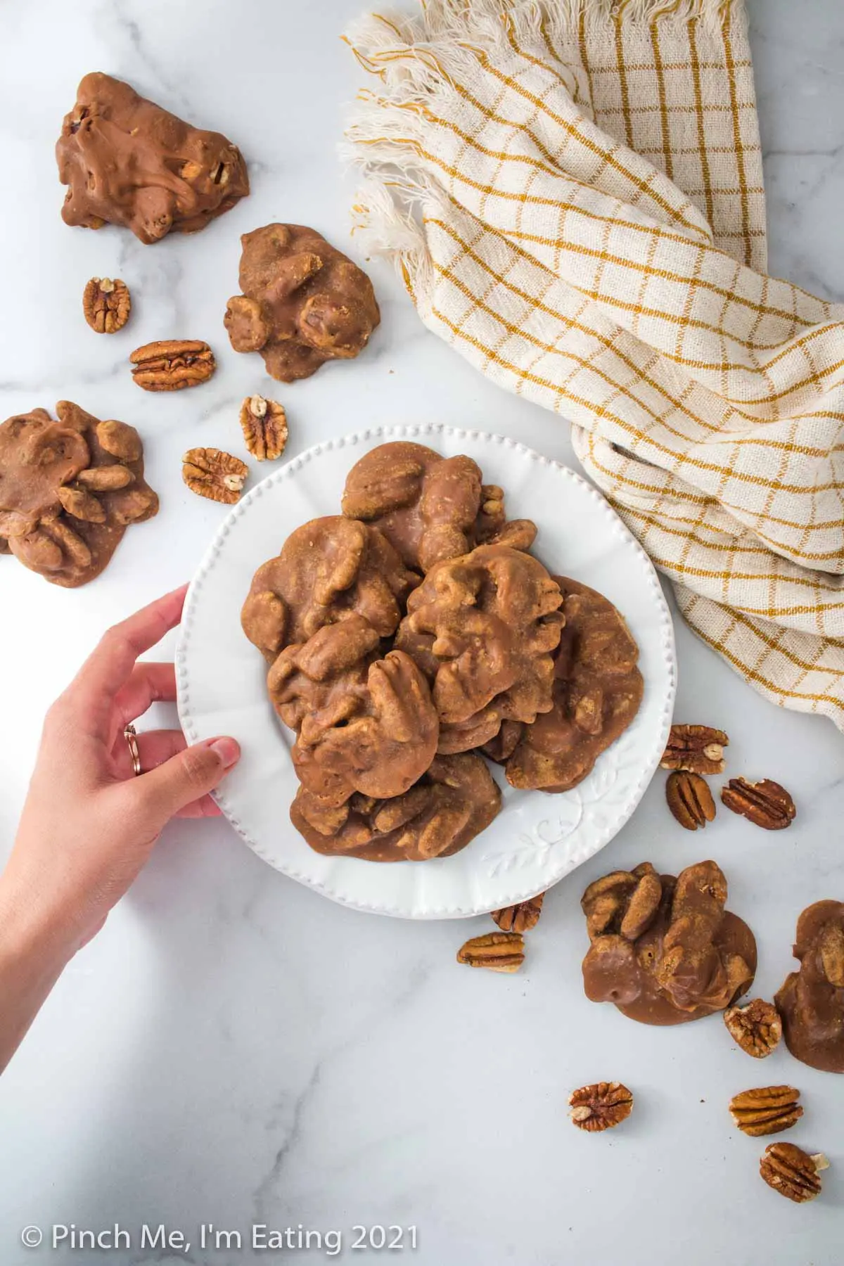 Overhead view of Southern pecan pralines on a white plate on a marble background. A hand is holding the plate, and pralines and pecans are scattered on the table around the plate.