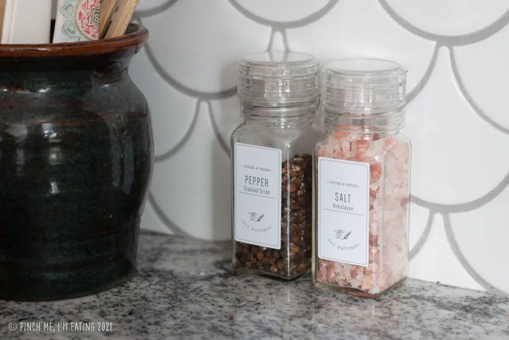 Pink Himalayan salt and black pepper in matching labeled clear spice grinders on countertop