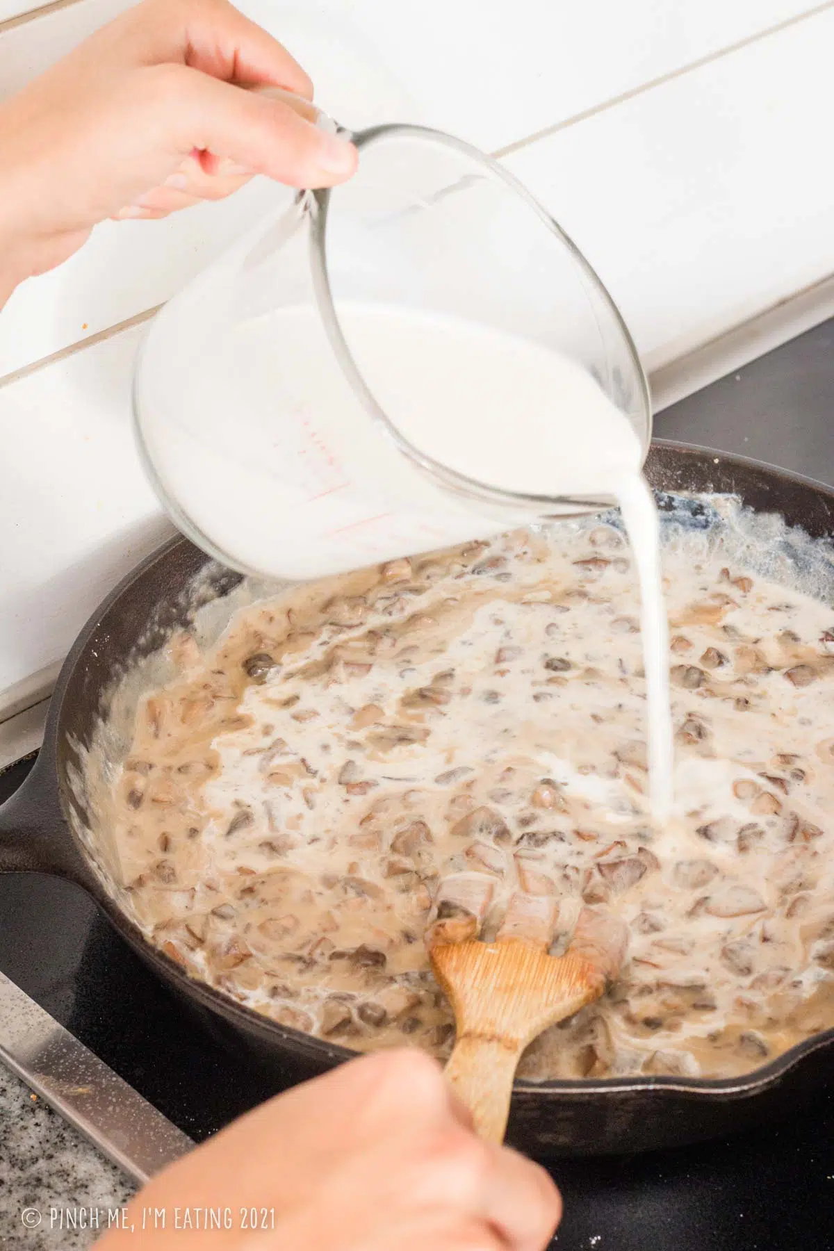 Hand pouring half-and-half into a cast iron skillet with creamy homemade mushroom soup.