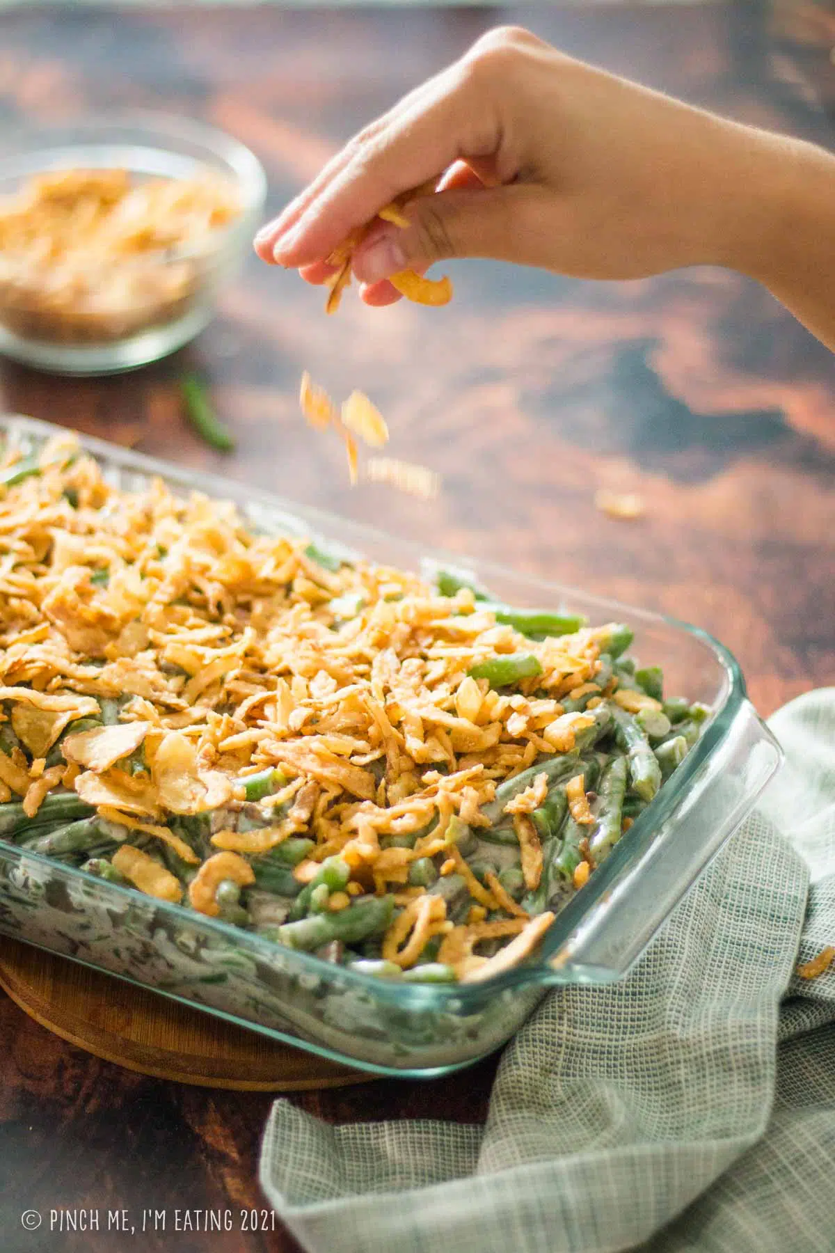 Hand sprinkling crispy fried onions over top of a green bean casserole in a glass baking dish.