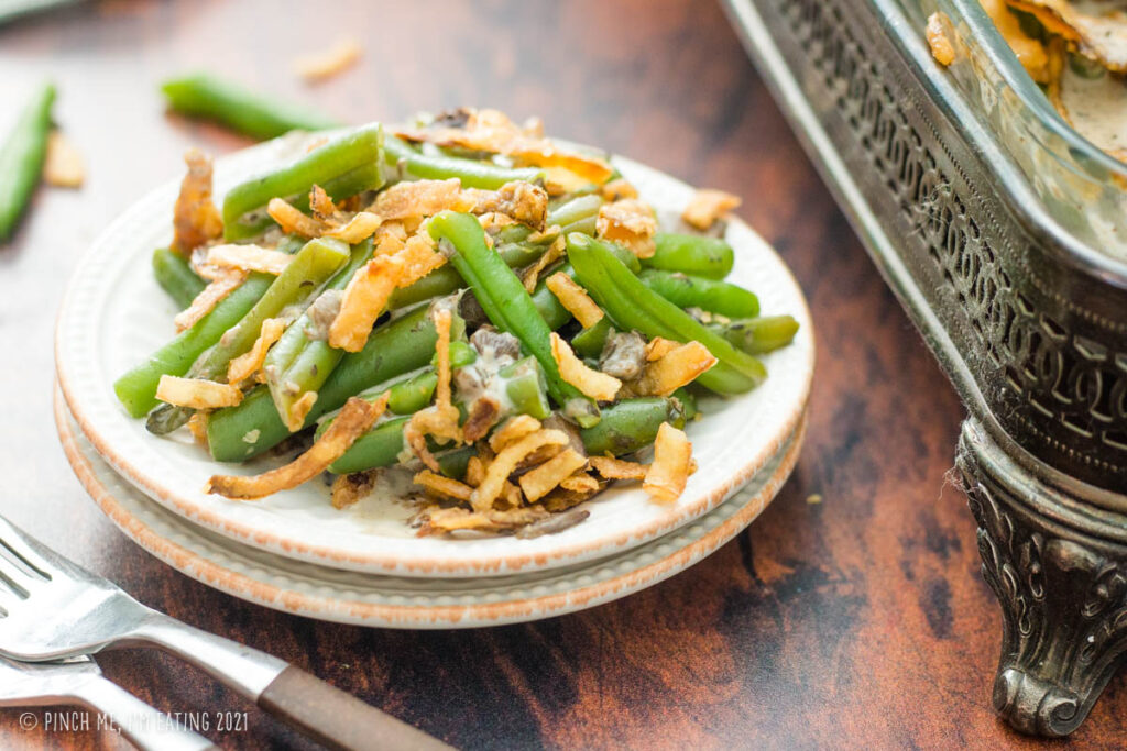 Fresh green bean casserole on a small white plate in front of a decorative casserole dish.