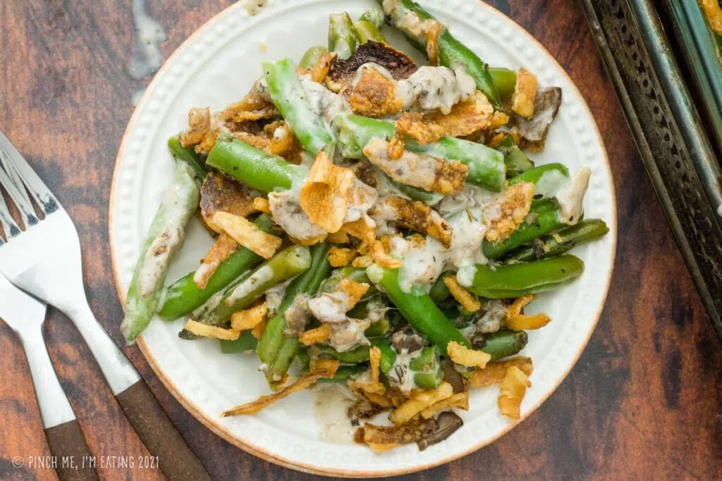 Fresh green bean casserole with crispy fried onions on a small white plate next to a decorative casserole dish.