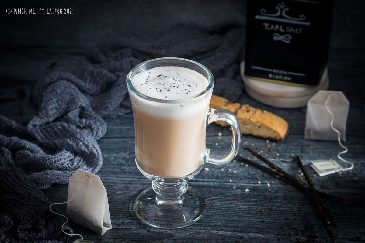 London Fog latte in a glass mug on a dark wood background with tea bag in front and biscotti behind it.