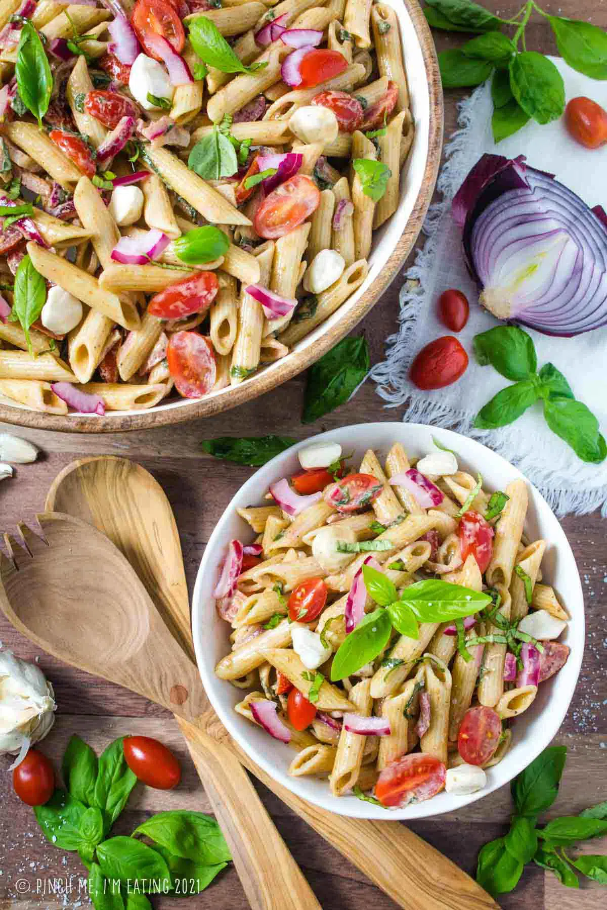 Overhead shot of creamy balsamic caprese pasta salad in a white bowl next to large wooden serving bowl.