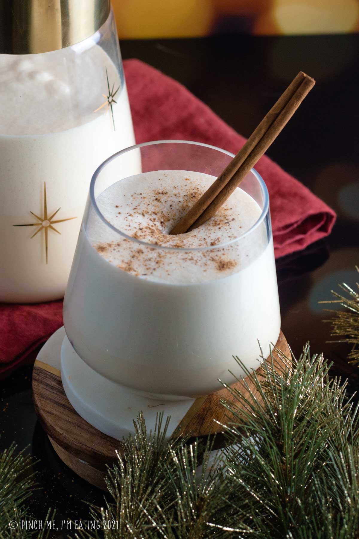 Glass of eggnog with cinnamon stick, topped with ground nutmeg, with pitcher in the background.