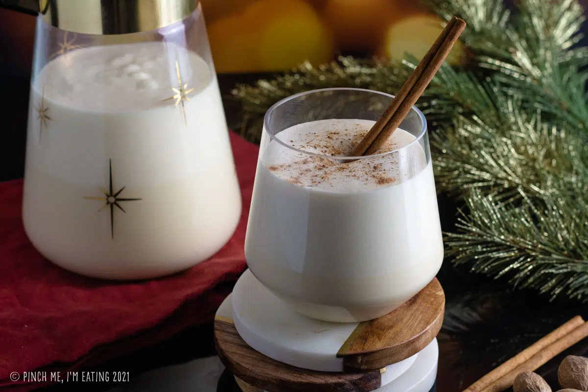 Glass of eggnog with cinnamon stick, topped with ground nutmeg, with pine branch and pitcher of eggnog in the background.