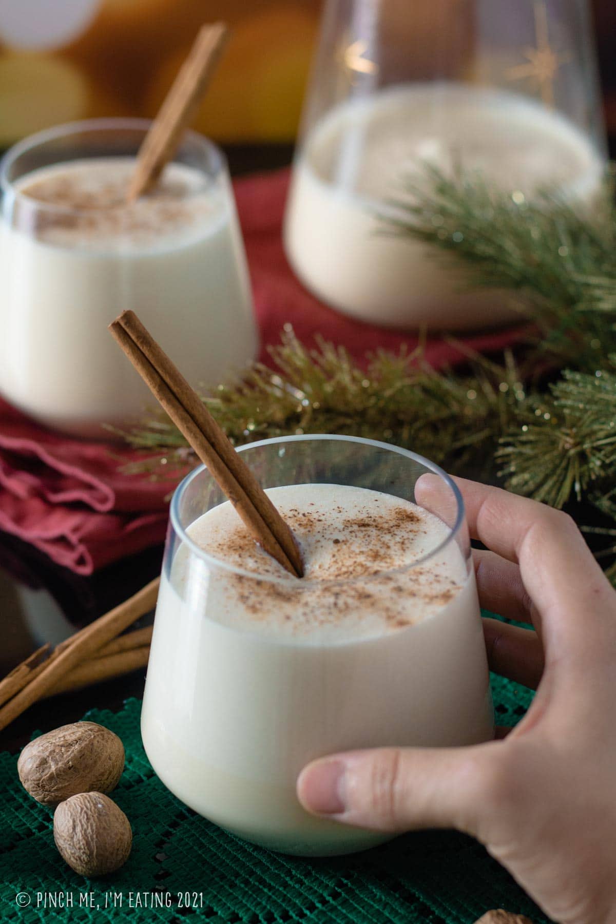 Hand holding glass of eggnog with cinnamon stick, topped with ground nutmeg.