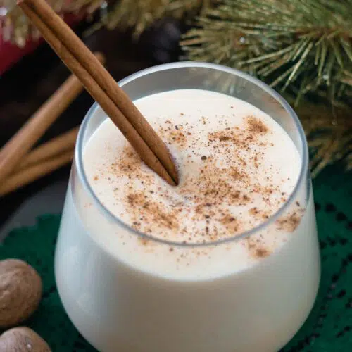 Glass of eggnog with cinnamon stick, topped with ground nutmeg.