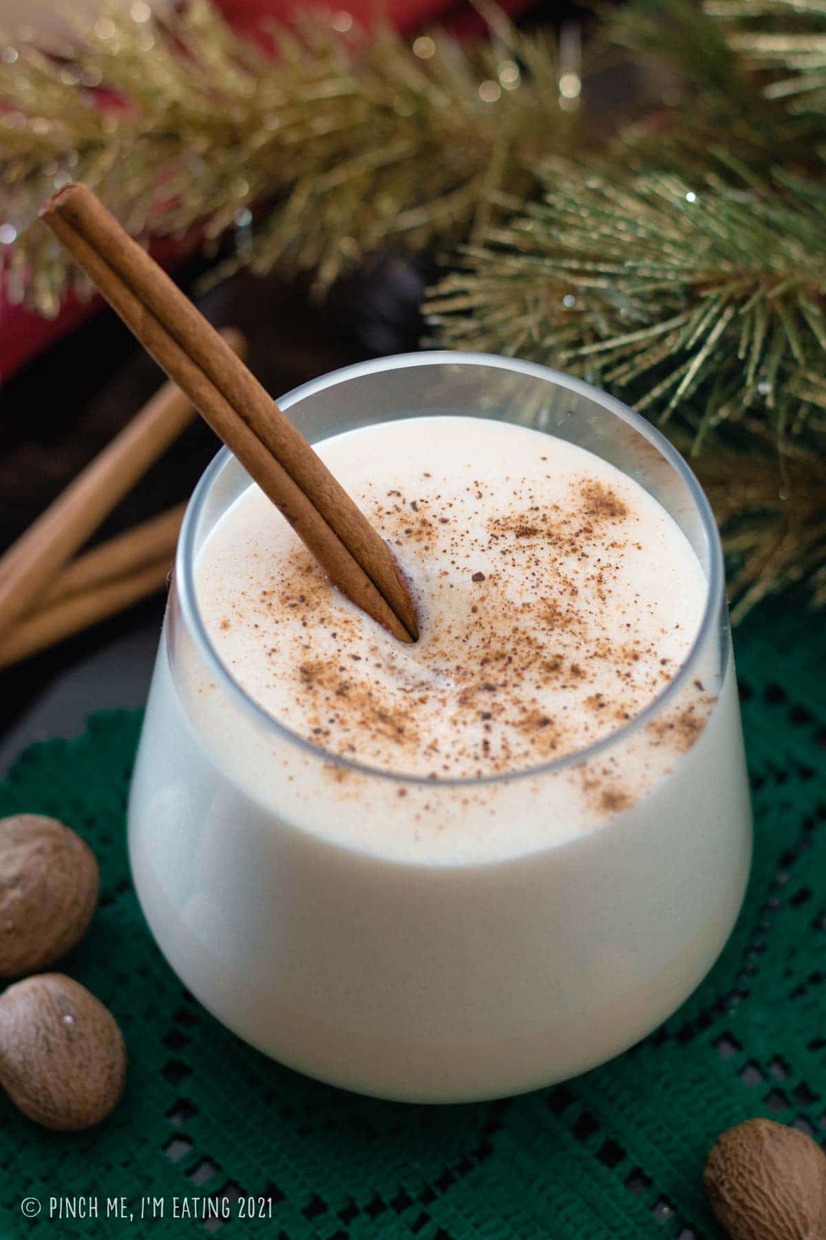 Glass of homemade eggnog with cinnamon stick, topped with ground nutmeg. Whole nutmeg is in the foreground and evergreen branch is in the background.