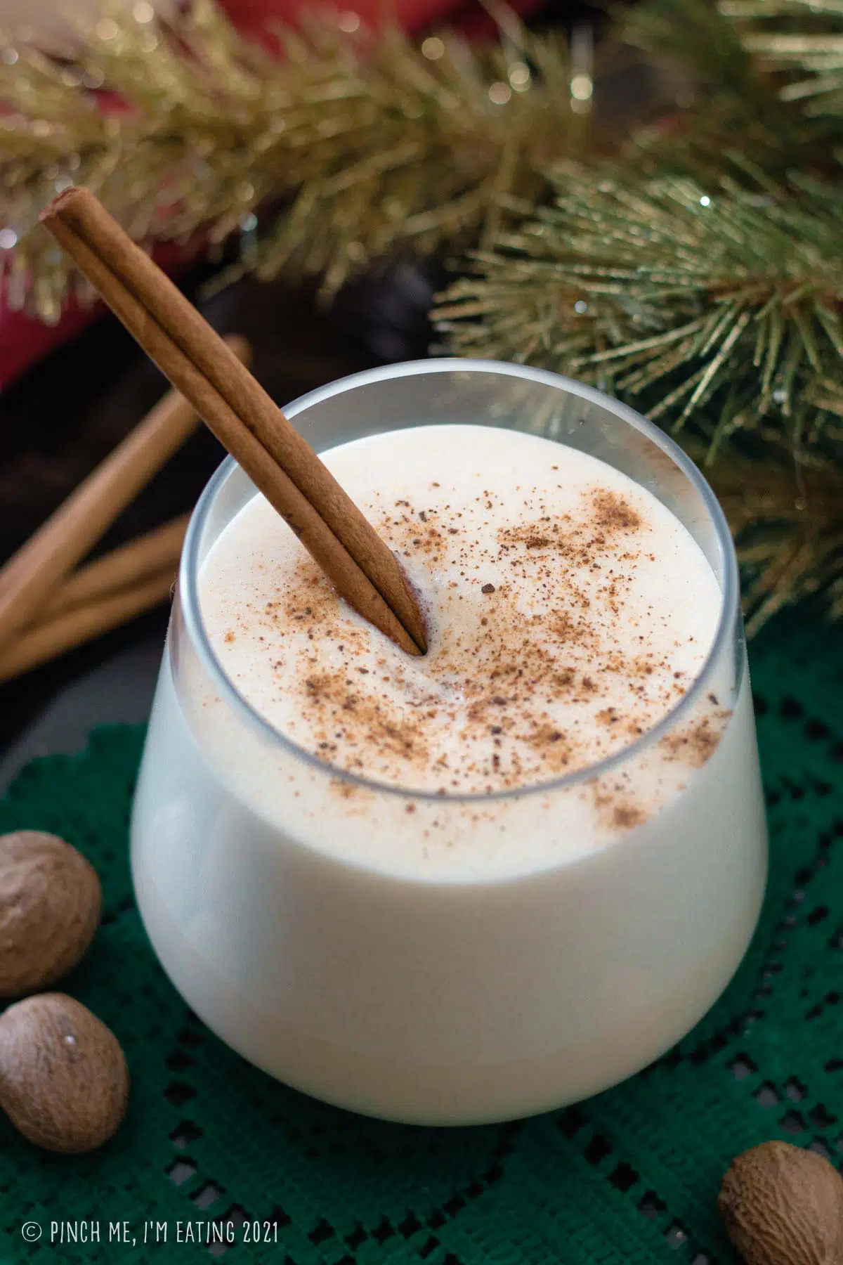 Glass of homemade eggnog with cinnamon stick, topped with ground nutmeg. Whole nutmeg is in the foreground and evergreen branch is in the background.