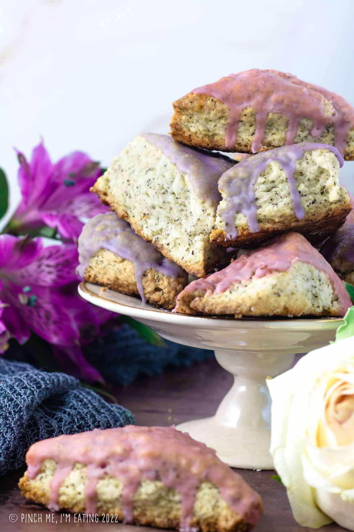 Earl grey scones with pink and purple lavender glaze stacked on a white cake stand with flowers in the background.