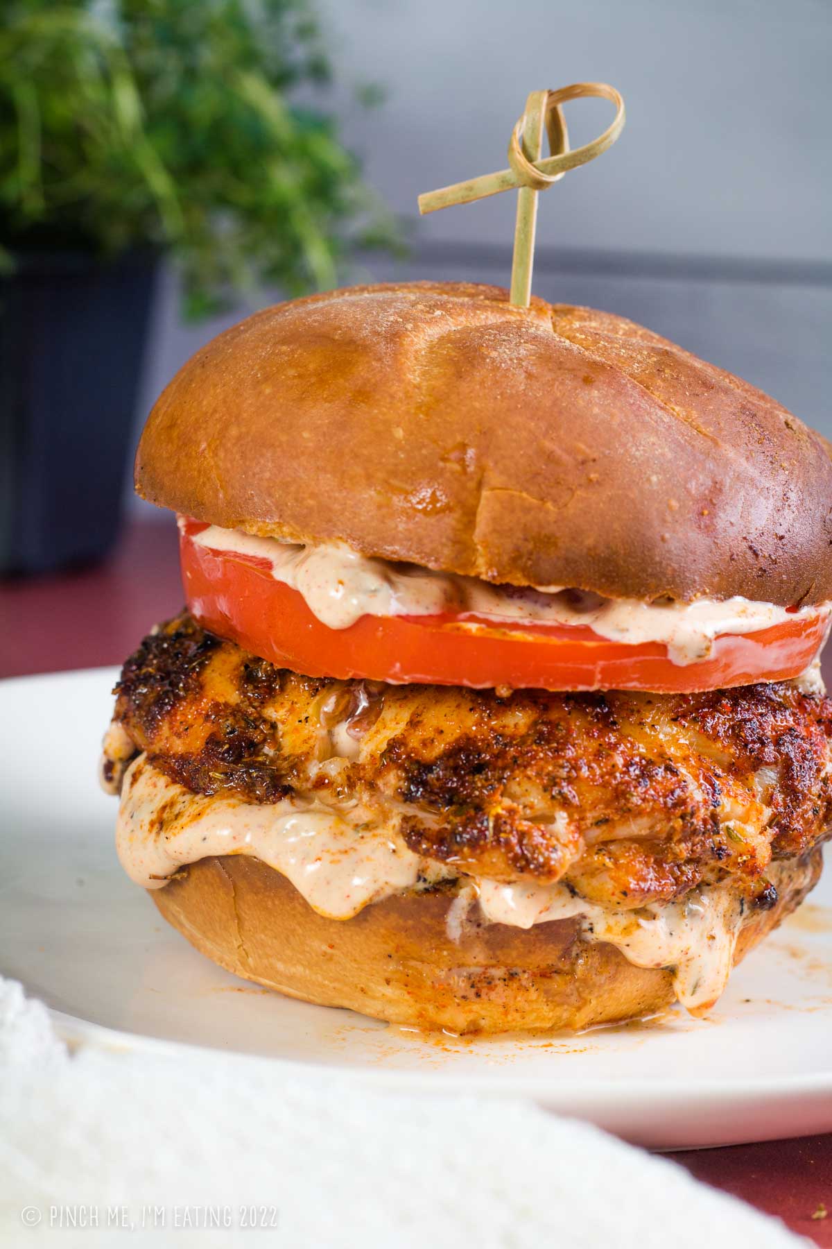 Close up of juicy blackened grouper sandwich with Cajun remoulade and a tomato slice on a white plate with a decorative toothpick.