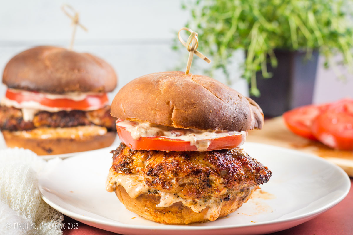 Two blackened grouper sandwichs with Cajun remoulade and tomato on  white plates with decorative toothpicks and a plant in the background.