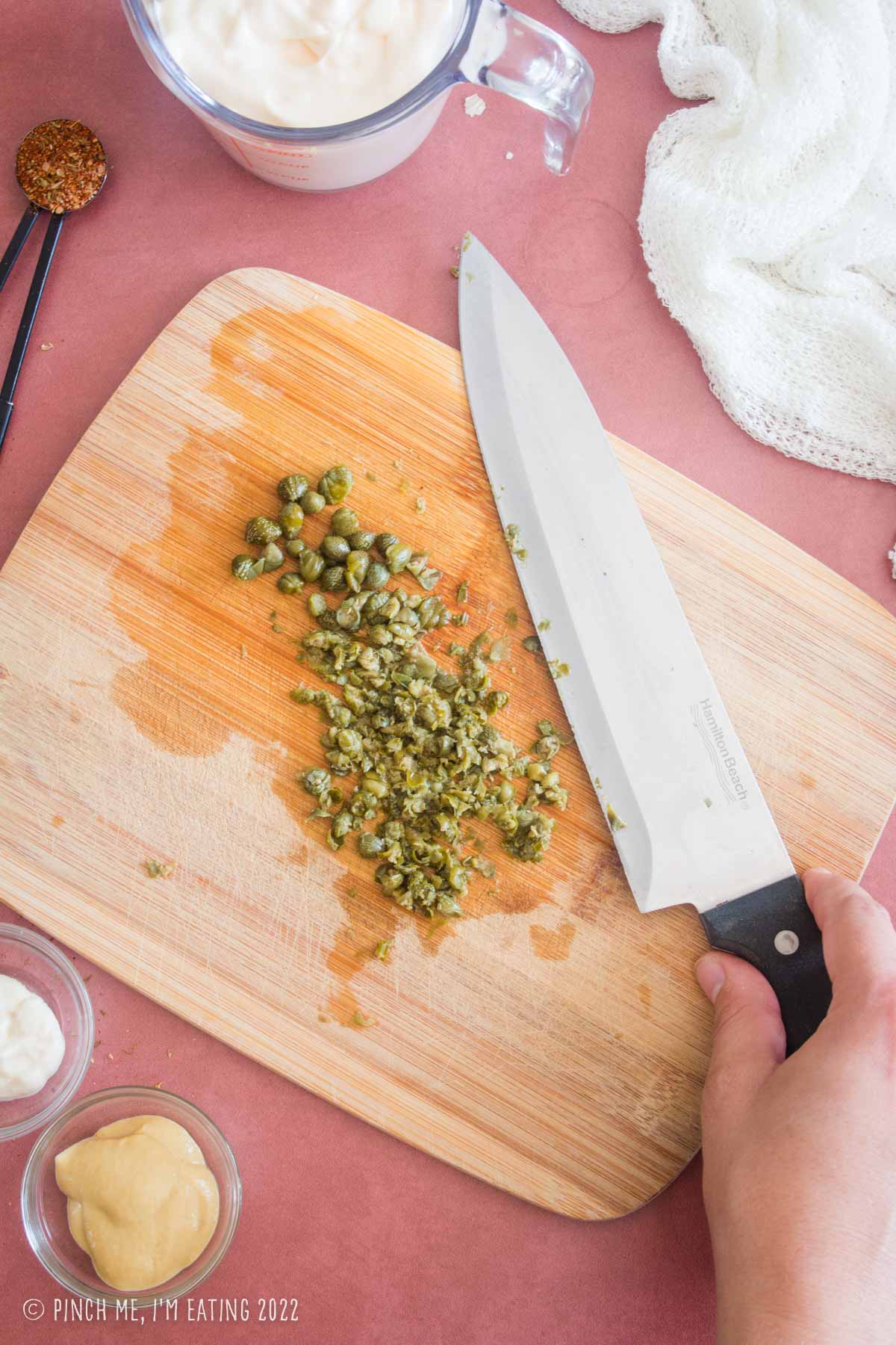 Minced capers on a wooden cutting board with a large chef's knife.
