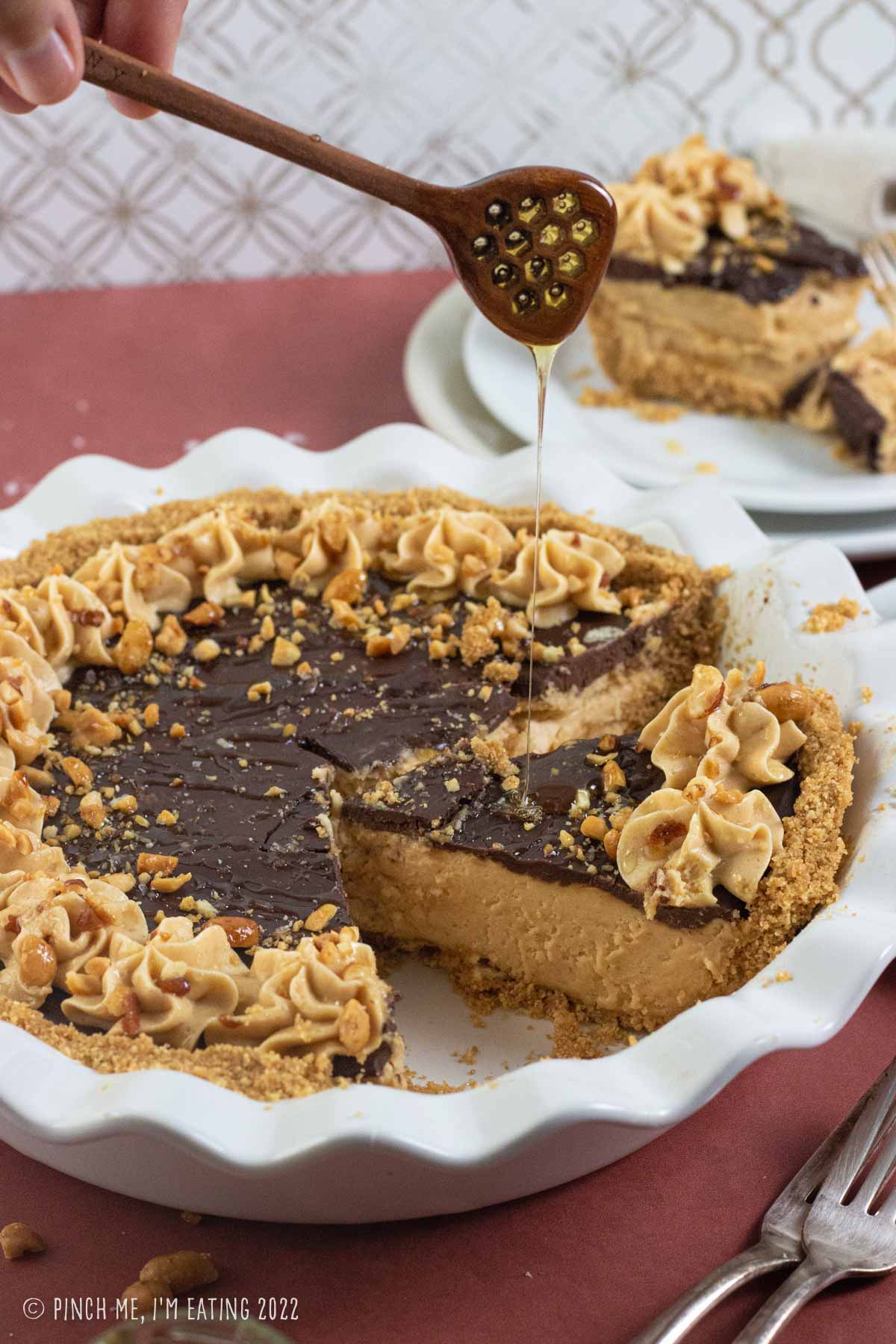 Honey drizzling on top of a peanut butter pie with graham cracker crust and chocolate ganache in a white pie plate.