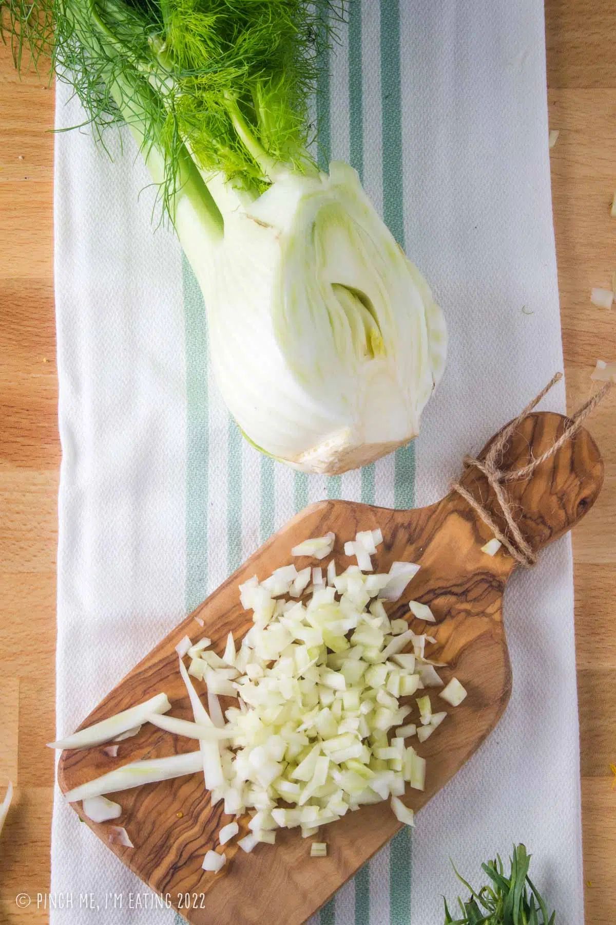 Overhead view of a bulb of fresh fennel on a kitchen towel above a cutting board with finely diced fennel bulb.