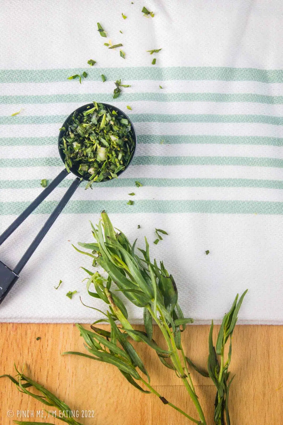 Chopped fresh tarragon in a measuring spoon on a kitchen towel on butcher block counter next to a stem of fresh tarragon leaves.
