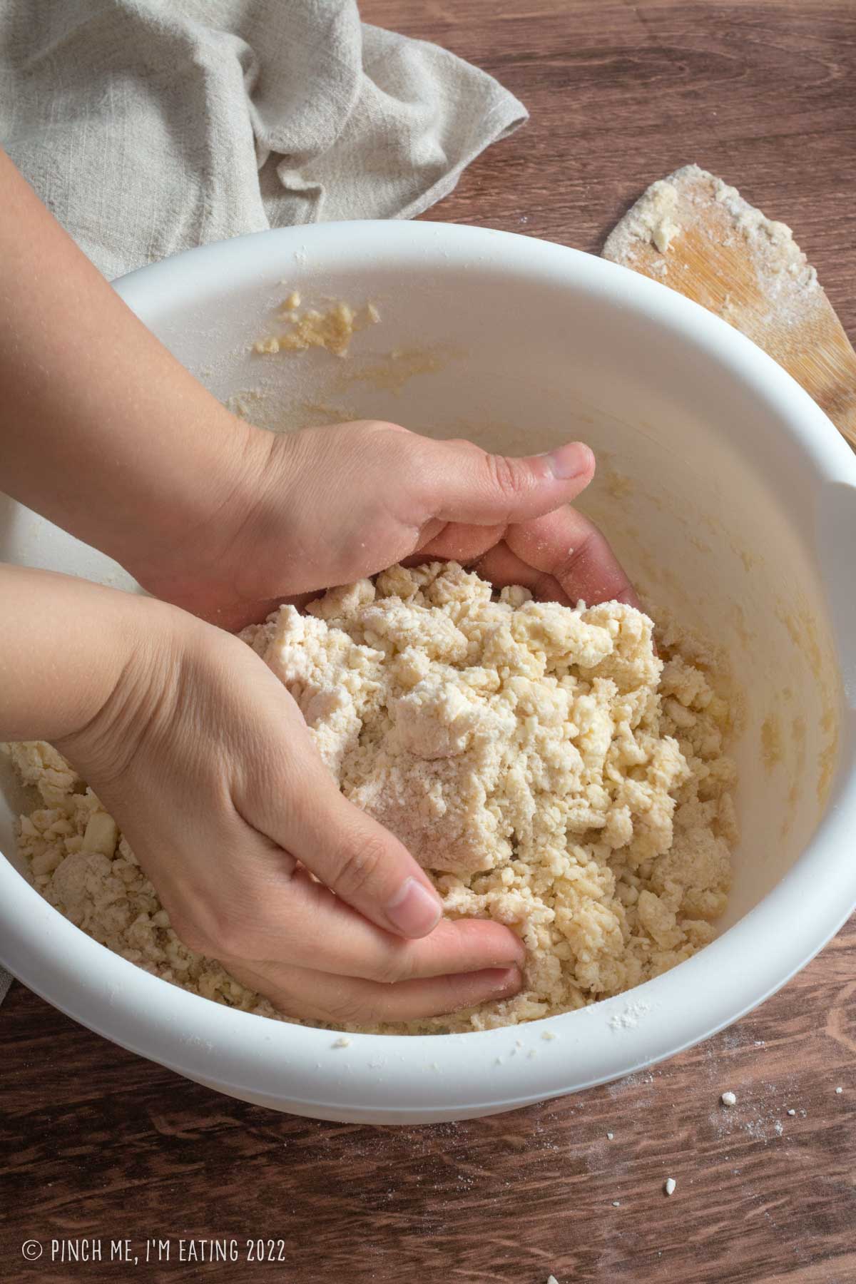 Hands patting together crumbly scone dough in a white bowl.