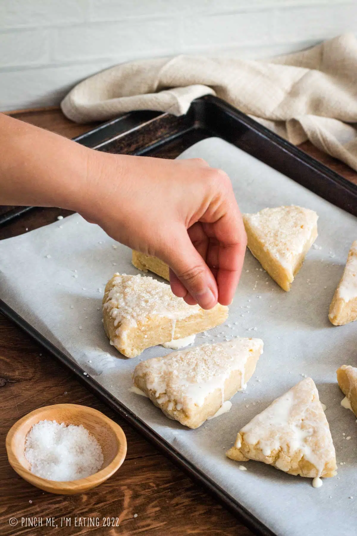 A hand sprinkling coarse sanding sugar onto plain scones brushed with heavy cream on a parchment-paper-lined baking sheet.
