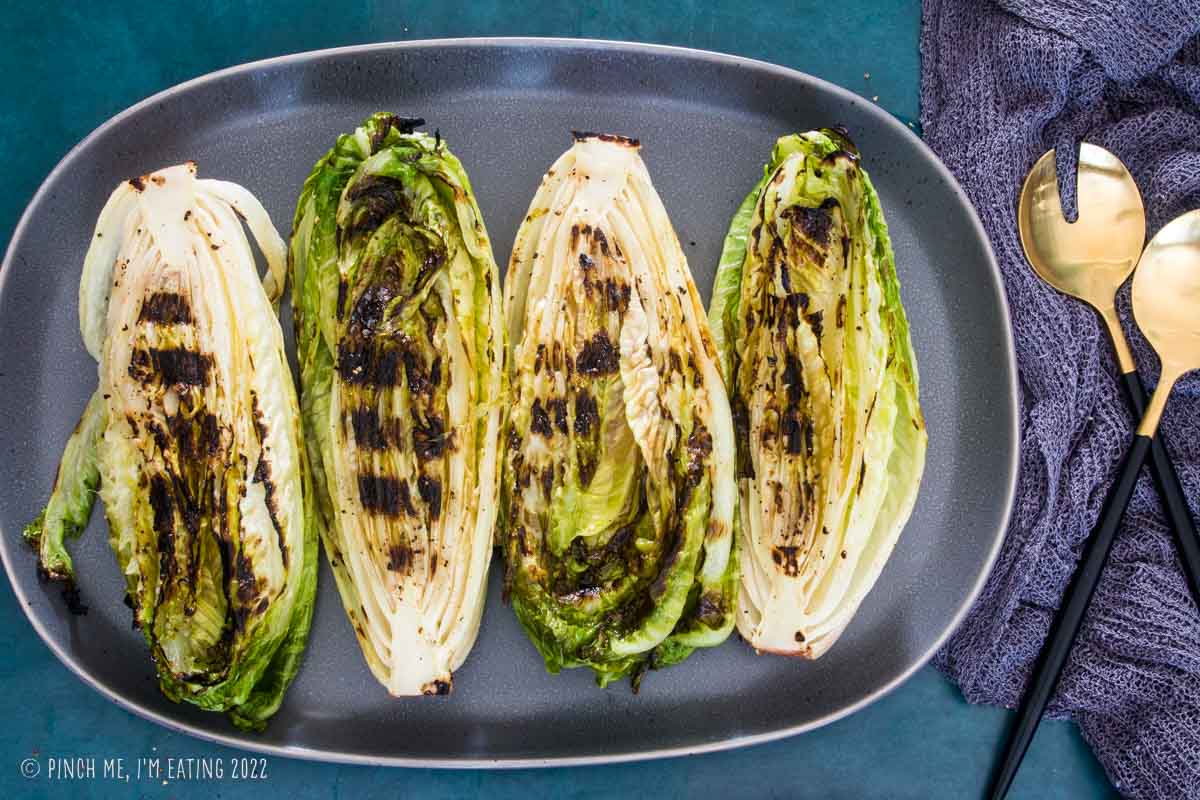 Grilled hearts of romaine on a gray platter with gold serving utensils.