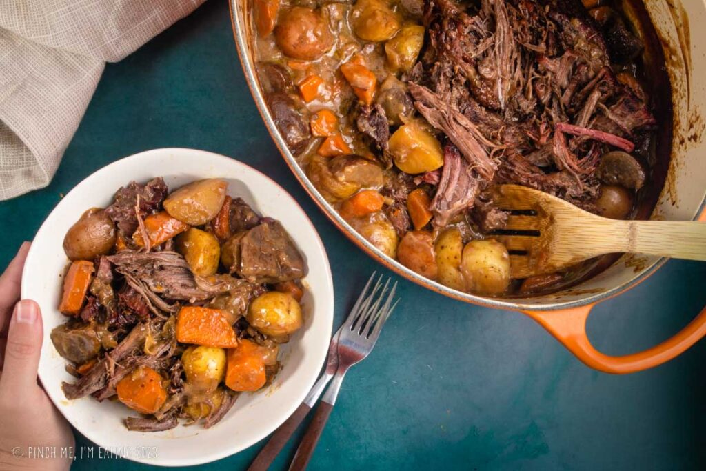White bowl of pot roast next to pot roast with carrots and potatoes in a Dutch oven.