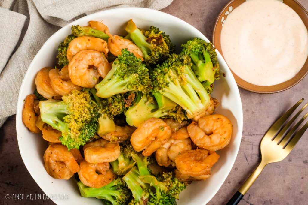 Overhead view of hibachi shrimp and broccoli in white bowl with yum yum sauce on the side.