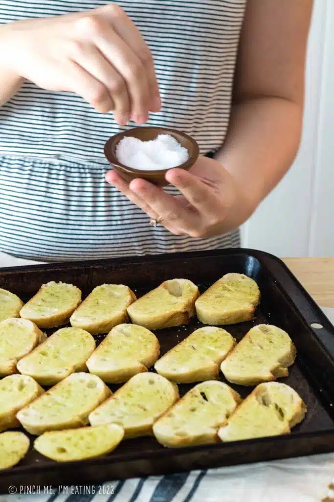 Sprinkling salt onto baguette slices on a baking sheet for making crostini.