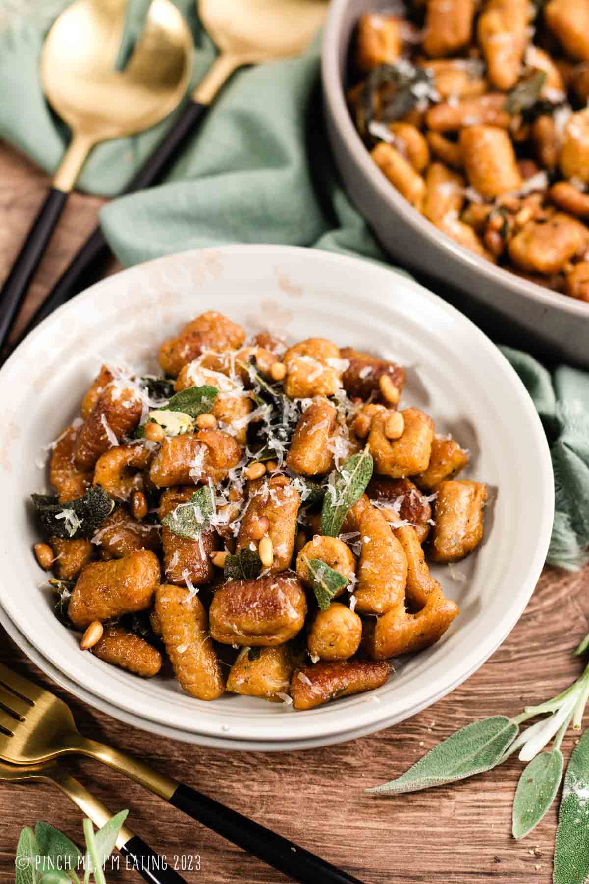 Homemade pumpkin gnocchi with crispy sage, pine nuts, and parmesan cheese in a bowl on a wood table with fresh sage leaves.